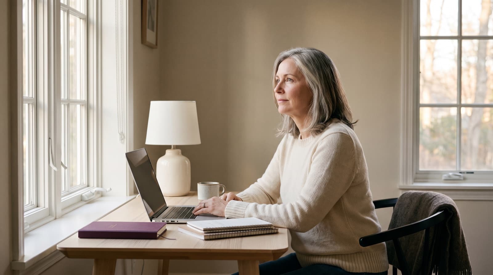 A woman in her early fifties pauses at her desk beside a sunlit window, looking away from her laptop.
